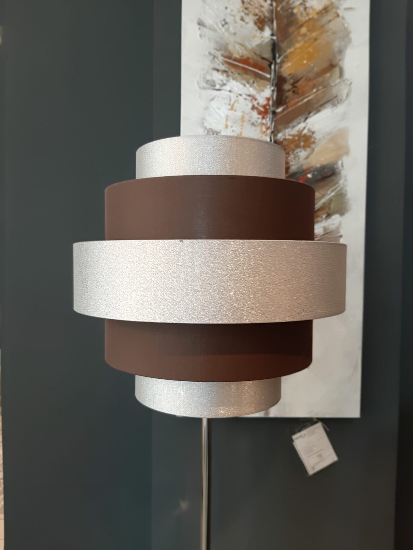 Smeđa i srebrna cilindrična lampa ispred apstraktne slike na tamnom zidu.